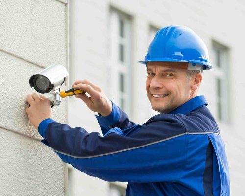 Technician wearing a blue uniform and hard hat installing a CCTV camera on a light-colored exterior wall of a Melbourne home or small business. Technician installing a CCTV camera